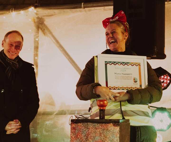 Two people at an event, one holding a certificate, festive setting with lights.